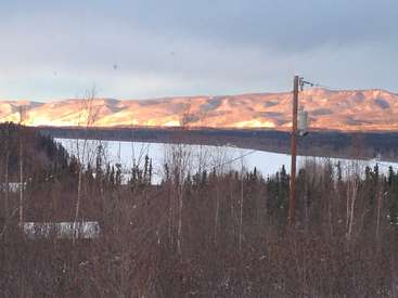 Das Bild zeigt eine ruhige Winterlandschaft mit einem zugefrorenen See, schneebedeckten Bergen und einem Telefonmast vor einer Kulisse aus Bäumen und einem bewölkten Himmel.