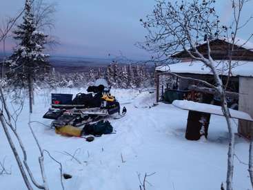 Das Bild zeigt ein Schneemobil, das vor einer Hütte geparkt ist, mit einer ruhigen Schneelandschaft und einem Gewässer im Hintergrund.