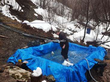 Das Bild zeigt eine Person, die in einem behelfsmäßigen Pool steht, umgeben von schneebedecktem Boden und Bäumen, mit einer blauen Plane, die die Oberfläche des Pools bedeckt.