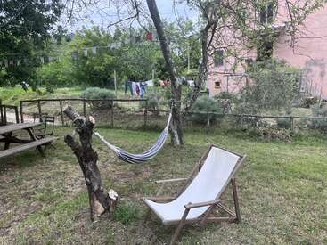 A peaceful backyard scene with a hammock strung between trees, a deck chair, picnic table, prayer flags, laundry hanging, and greenery surrounding a pink building.