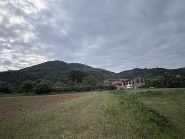 A cloudy sky hovers above green hills and a rural field, with a few buildings and trees in the distance, creating a peaceful countryside scene.