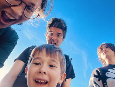 Four people pose for a fun, playful selfie against a bright blue sky. They are smiling, laughing, and making silly faces, enjoying a joyful moment together.