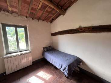 This small room features a single bed with a gray cover, rustic wooden beams, a tiled ceiling, a window with natural light, and white plaster walls.
