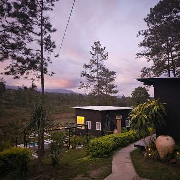 Un paisaje sereno al atardecer con modernas cabañas negras, exuberante vegetación, altos pinos, un sinuoso sendero y montañas bajo un suave cielo vespertino de colores pastel.