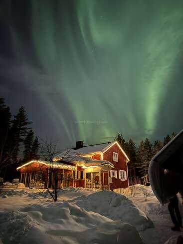 Uma aconchegante cabana vermelha, com uma iluminação acolhedora, fica no meio da neve profunda e dos pinheiros. No céu, a deslumbrante aurora boreal verde dança em uma noite mágica de inverno, repleta de estrelas.