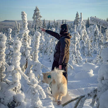 Uma pessoa vestida com roupas de inverno está em uma floresta coberta de neve, apontando para a frente. Ao lado dela está um cachorro fofinho, cercado por árvores cobertas de gelo sob um céu azul claro.