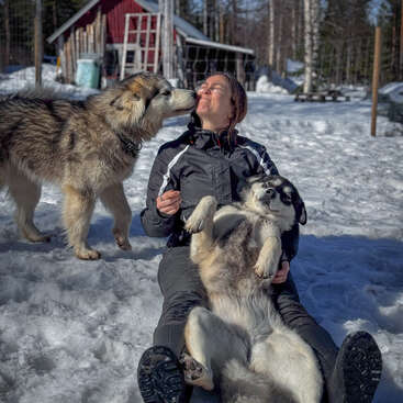 Eine Person sitzt im Schnee und hält einen entspannten Hund im Arm, während ein anderer Hund ihr liebevoll das Gesicht leckt. Eine rote Hütte und verschneite Bäume bilden den Hintergrund.