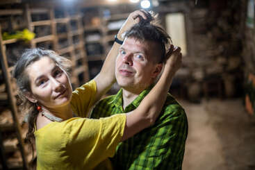 A woman in a yellow shirt playfully pulls a man's hair in a rustic indoor setting. Both appear amused, creating a lighthearted, candid atmosphere together.