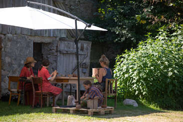 Four people, including a child, sit outside under a large umbrella, working with clay at a table near a rustic shed, surrounded by greenery and sunlight.