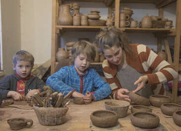 A woman and two young boys make pottery together in a cozy studio, surrounded by handmade clay bowls, tools, and shelves filled with finished ceramic pieces.