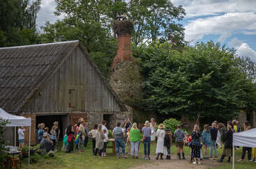 A group of people gathers outdoors near an old wooden barn, listening to someone speak. On a tall chimney, a large stork nest is visible above.