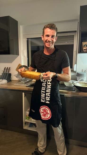 A smiling man stands in a kitchen, wearing an Eintracht Frankfurt apron. He proudly holds a freshly baked dish, radiating happiness and satisfaction with his cooking.