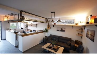 This cozy, modern room features a small bar, comfortable gray sofa, wall shelves, a guitar, hanging lights, plants, and scenic framed photographs on the walls.