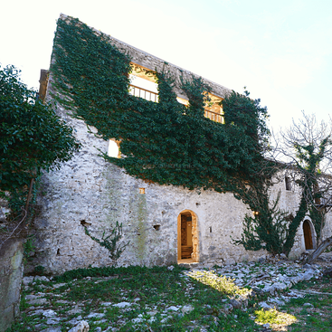 Cette image montre un vieux bâtiment en pierre partiellement recouvert de lierre vert. La lumière du soleil illumine une porte ouverte. Le sol est rocailleux, parsemé de touches de verdure.