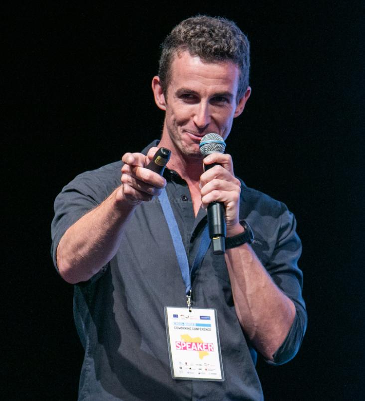 A man with curly hair, wearing a dark shirt, confidently speaks into a microphone on stage, pointing at the audience, wearing a conference badge labeled "SPEAKER".