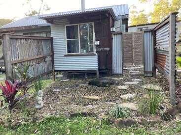 Una casita rústica con paredes y tejado de chapa ondulada, rodeada de un sencillo jardín, escalones, plantas, una estatua de Buda y vallas de madera y metal.