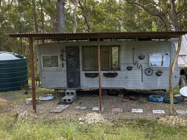 Una caravana rústica y decorada se asienta bajo un tejado metálico en una zona boscosa. El suelo está cubierto de baldosas artísticas y cerca hay un gran depósito de agua.
