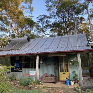Una casita rústica con tejado de chapa ondulada y calentador de agua solar, rodeada de árboles, plantas, una acogedora decoración de madera y una acogedora puerta amarilla.