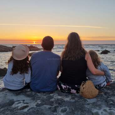 L'image représente une famille de quatre personnes assise sur une plage rocheuse, contemplant le coucher de soleil sur l'océan, le soleil se couchant à l'horizon.