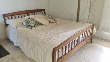 Ein gemütliches Schlafzimmer mit einem Holzbettgestell, cremefarbener Bettwäsche und drei dekorativen Kissen. Das Zimmer hat weiße Wände und ein rustikales, leicht abgenutztes Aussehen.