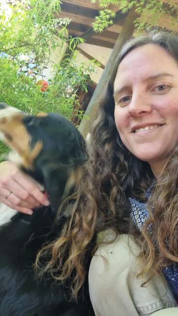 Eine Frau mit langen lockigen Haaren lächelt in die Kamera und streichelt einen schwarz-braun-weißen Hund. Hinter ihnen sind Grünpflanzen und hölzerne Dachbalken zu sehen.