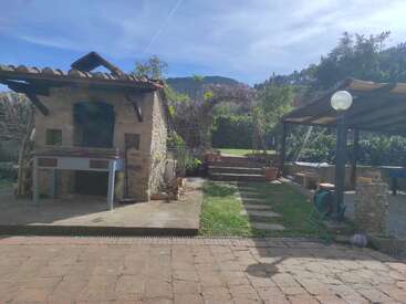 La imagen muestra una cocina rústica al aire libre con un horno de piedra, un patio, una zona de descanso cubierta, unos escalones que dan al jardín, vegetación y montañas al fondo, bajo un cielo azul.