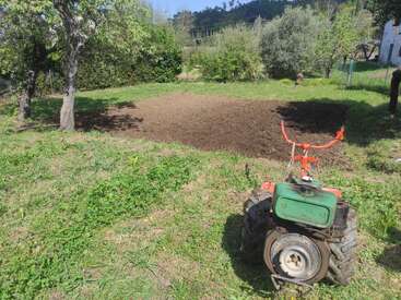 Una parcela de jardín recién labrada está rodeada de césped, árboles y arbustos. En primer plano se ve una pequeña motocultivadora de color verde y naranja.