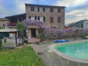 Una casa rústica de piedra con flores trepadoras de color púrpura, una piscina, vegetación y una bandera al fondo, bajo un cielo parcialmente nublado con vistas al campo.