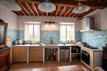 Esta cocina rústica cuenta con armarios de madera, vigas de madera a la vista, dos ventanas, azulejos de color azul para el salpicadero, electrodomésticos modernos, estanterías abiertas, una iluminación luminosa y un ambiente acogedor y agradable.