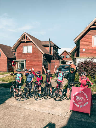 Six cyclists pose cheerfully with bikes in front of wooden cabins on a sunny day. A pink sign reads "Gravel Travel, Pto Varas, Chile."