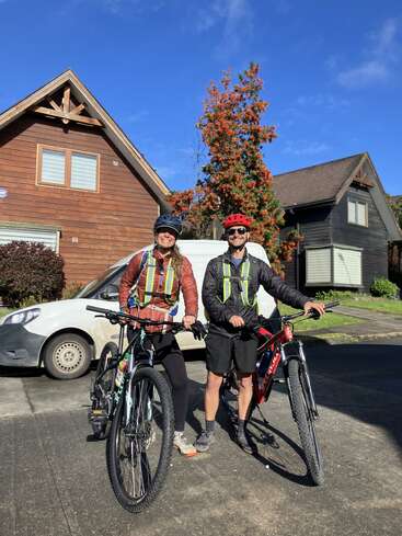 Two people in helmets and reflective vests pose with their bicycles outside wooden houses on a sunny day. A white van and colorful tree are behind them.