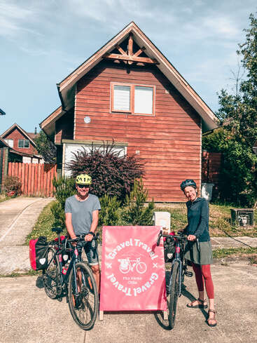 Two cyclists with helmets stand by their bicycles in front of a wooden house. A pink sign reads “Gravel Travel Pto Varas Chile.” Sunny, cheerful scene.