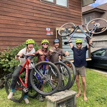 Four happy people wearing helmets pose with bicycles outside a wooden house. One lifts a bike overhead, all smiling, radiating excitement and camaraderie on a grassy lawn.