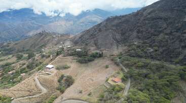 This aerial image shows a mountainous landscape with winding roads, scattered houses, green patches of trees and vegetation, cloudy skies, and expansive distant mountain ranges in view.