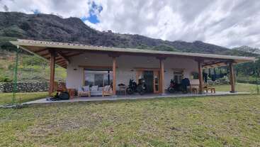 A modern house with a large covered patio, outdoor seating, and two motorcycles. It sits on a grassy yard, backed by hills and cloudy skies.