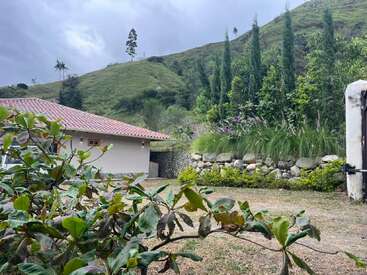 A peaceful rural scene with a house having a red-tiled roof, surrounded by lush greenery, hills, stone wall, tall trees, and overcast sky above.