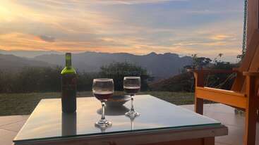 Two glasses of red wine and a bottle sit on a glass table outdoors, overlooking serene mountains at sunset, with a wooden chair completing the peaceful view.