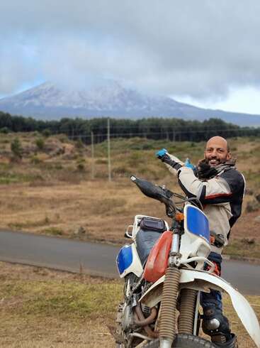 A smiling man in motorcycle gear stands beside his bike, pointing happily toward a distant, cloud-covered mountain in a scenic, grassy landscape with trees.