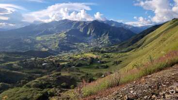 This image showcases a stunning mountain landscape with rolling green hills, scattered houses, lush valleys, dramatic shadows, and fluffy clouds under a bright blue sky. Serene scenery.