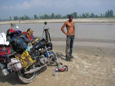 A shirtless man stands on a sandy riverbank beside a heavily loaded motorcycle. The scene is adventurous, with distant water, trees, and a blue sky.