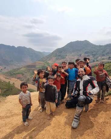 A joyful group of children and a man in motorcycle gear pose together on a scenic hillside, smiling and waving with mountains and terraced fields in the background.