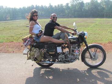 A smiling man and woman ride a Royal Enfield motorcycle on a rural road. Both wear sunglasses, enjoying the breeze, with lush greenery in the background.