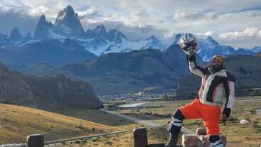 A man in motorcycle gear poses triumphantly, holding his helmet aloft against a stunning mountain backdrop, with a small town and winding road below, under dramatic clouds.