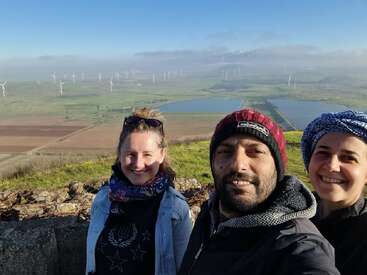 Three people smile for a selfie on a hillside. Behind them are fields, ponds, wind turbines, and distant mountains, under a partly cloudy blue sky.
