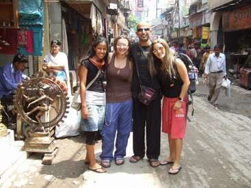 Four friends pose together in a busy street market, surrounded by shops, sculptures, and locals. They smile brightly, capturing a moment of travel and adventure.