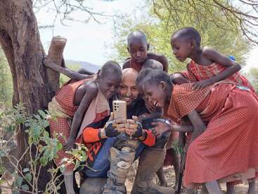 Un groupe d'enfants souriants, vêtus de vêtements à motifs, se rassemble avec enthousiasme autour d'un homme tenant un smartphone ; tous s'amusent joyeusement, assis en plein air sous un arbre.