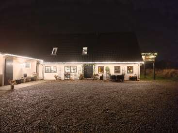 Ein gemütliches weißes Haus wird nachts von Lichterketten entlang des Daches beleuchtet. Topfpflanzen und Gartenmöbel schmücken den Kieshof davor.