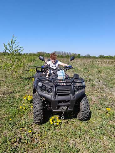 Eine Person mit lockigem Haar fährt an einem sonnigen Tag unter strahlend blauem Himmel mit einem Quad durch eine Wiese, umgeben von gelben Wildblumen.