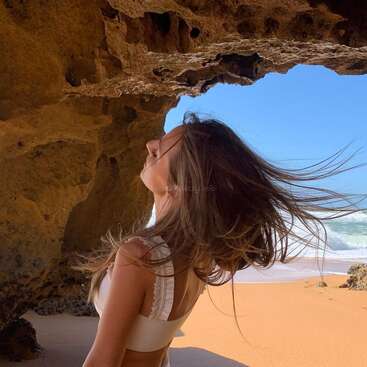 A woman with long hair stands in front of a cave on a sandy beach, with the ocean and blue sky visible in the background.