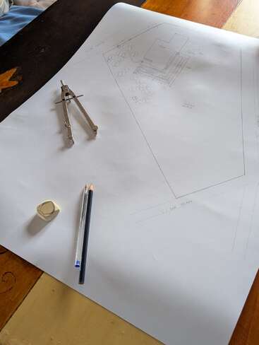 Ein großes Blatt Papier mit einem Gebäude- und Landschaftsplan, ein Metallzirkel, ein Bleistift, ein Kugelschreiber und ein Radiergummi werden auf einen Holztisch gelegt.